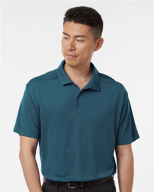 A man wearing a short-sleeve polo shirt in a muted teal blue color, paired with black pants and a black belt, photographed against a light gray studio background.